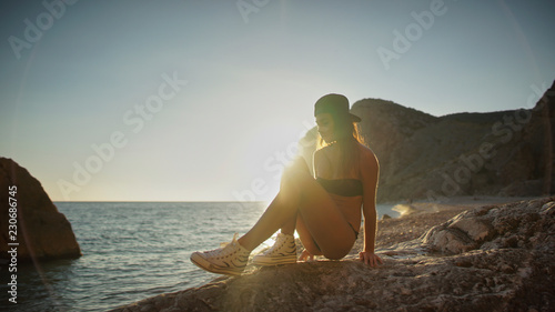 Beutiful young tnager woman seats on the rock in swimwear, cap and shouses against sunset