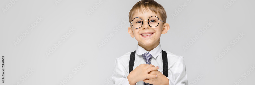 A charming boy in a white shirt, suspenders, a tie and light jeans stands on a gray background. A boy smiles and holds his hand tie
