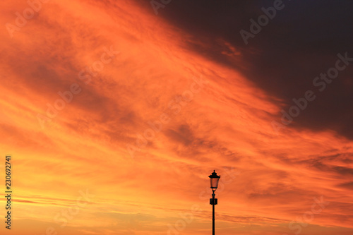 Sunset Sky and Vibrant Orange Cloudscape with Silhouette of One Vintage Lantern on City Street. Scenic Empty Sky Background at Sunset or Sunrise, Dusk and Dawn Beautiful Skyline Over the City View