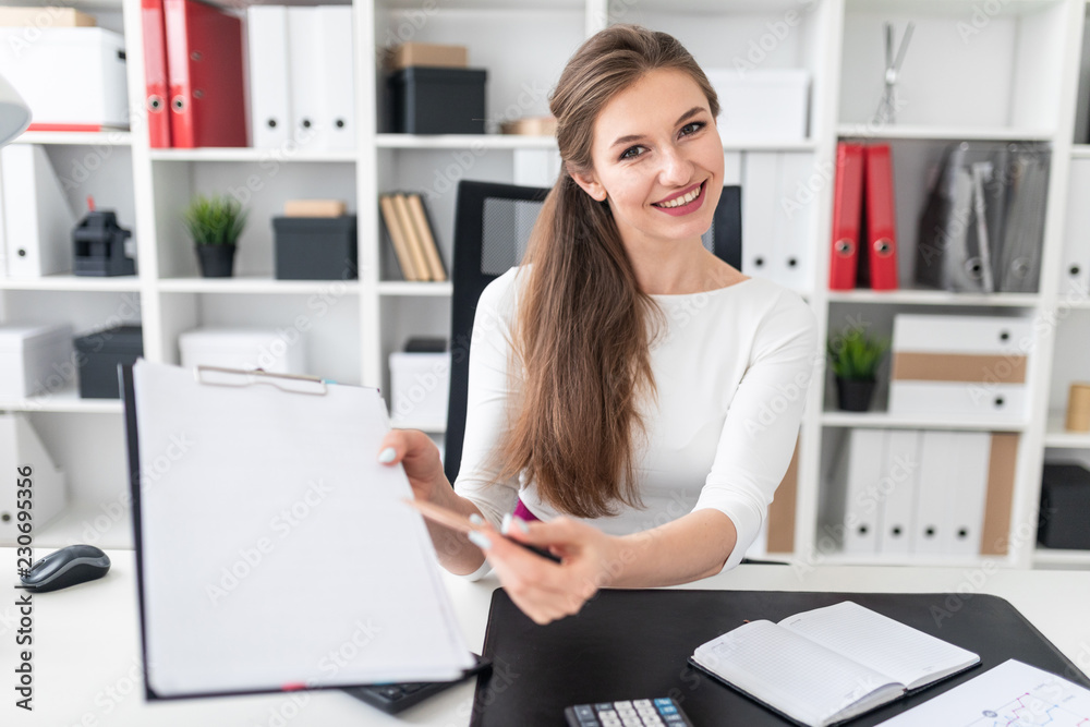 A young girl sitting at a table and shows a pen on an empty sheet.