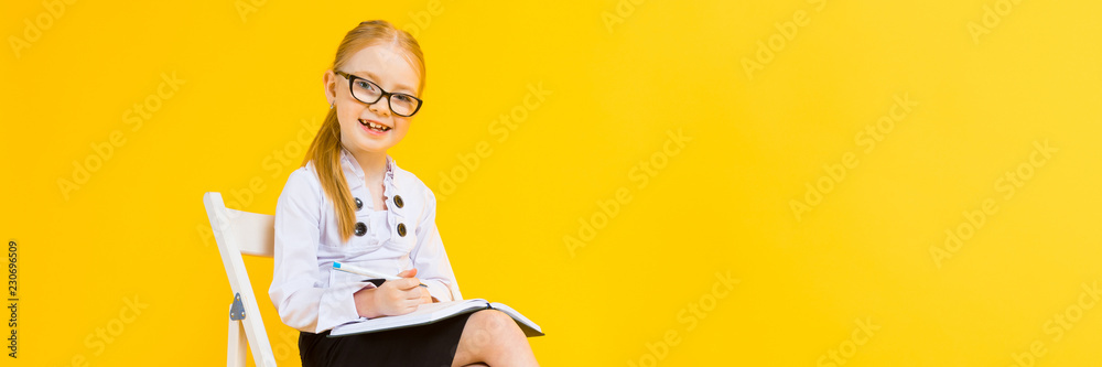 Girl with red hair on a yellow background. A charming girl in transparent glasses sits on a white chair and makes notes in a notebook. - obrazy, fototapety, plakaty 