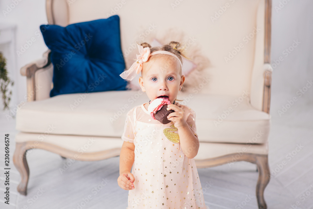 children's birthday. funny two-year-old Caucasian girl in pink dress standing to bedroom of house the background of couch and eating, holding cake dessert. Face and mouth smeared, smeared with cream children's birthday. funny two-year-old Caucasian girl in pink dress standing to bedroom of house the background of couch and eating, holding cake dessert. Face and mouth smeared, smeared with cream