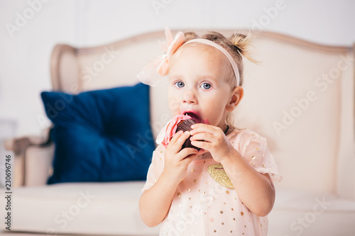 children's birthday. funny two-year-old Caucasian girl in pink dress standing to bedroom of house the background of couch and eating, holding cake dessert. Face and mouth smeared, smeared with cream photo