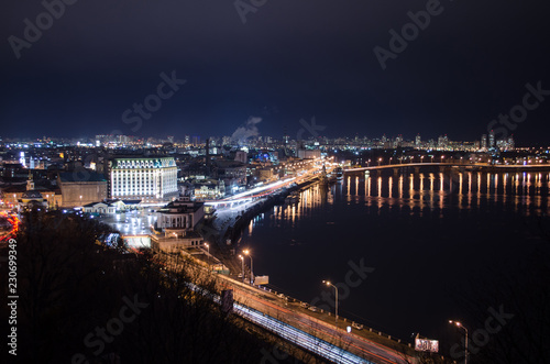 Panorama of night city landscape with river and bridge. Reflection of glowing lanterns in water.