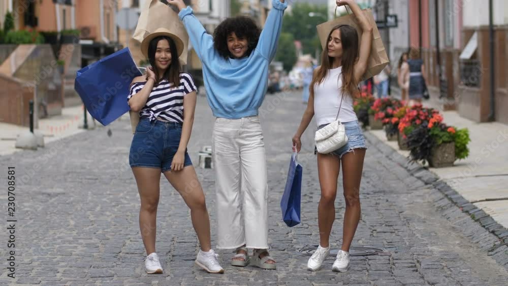 Lovely excited multi ethnic young female friends with shopping bags expressing joy, happiness and positivity, posing on shopping street over colorful urbanscape background. Lovely excited multi ethnic young female friends with shopping bags expressing joy, happiness and positivity, posing on shopping street over colorful urbanscape background.