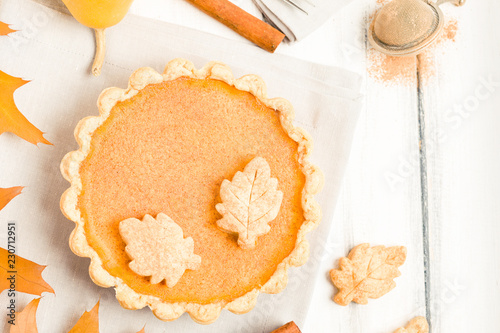 Pumpkin pie with cinnamon and cookies on gray napkins on white wooden background with autumn yellow leaves.