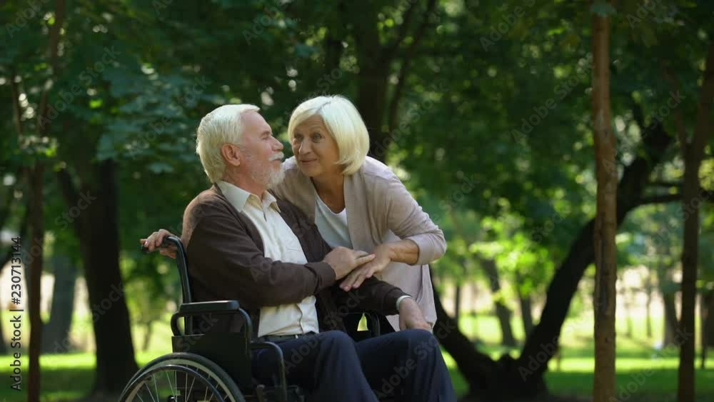 Mature couple resting in park looking at grandkids, man sitting in wheelchair Mature couple resting in park looking at grandkids, man sitting in wheelchair