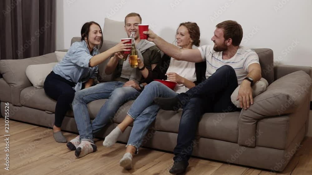 Friends are gathering together and having fun at the living room with loft interior. Male and female company, The girl with a big red bowl with popcorn, everyone drinking beer or soda, laughing Friends are gathering together and having fun at the living room with loft interior. Male and female company, The girl with a big red bowl with popcorn, everyone drinking beer or soda, laughing