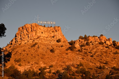 Low angle view of the city of Trinidad Colorado landmark sign with an early sunrise glow against the rocky butte and sky.