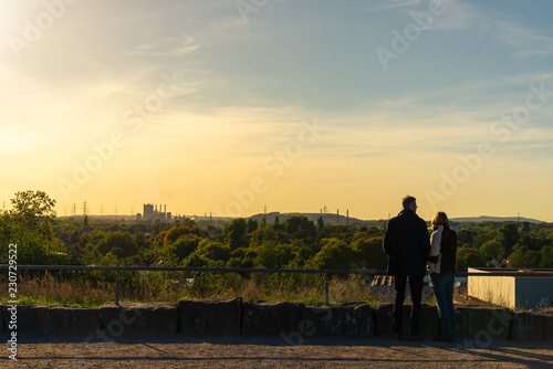 Scenic panoramic silhouette couple stand together and enjoy view of park and industrial area from the hill during sunset time at Europator Park in Gelsenkirchen, Germany. 
