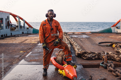 Seaman AB or Bosun on deck of offshore vessel or ship , wearing PPE personal protective equipment - helmet, coverall, lifejacket, goggles. He holds hammer photo