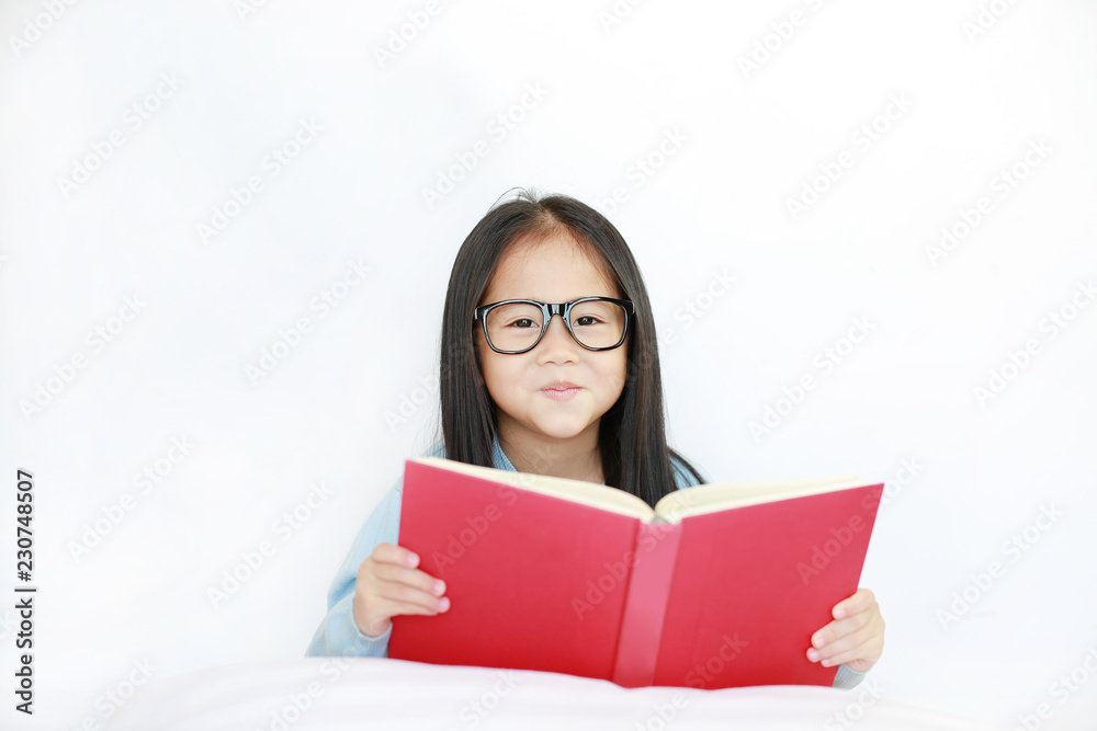 Beautiful happy little Asian kid girl reading hardcover book lying on bed against white background. Beautiful happy little Asian kid girl reading hardcover book lying on bed against white background.