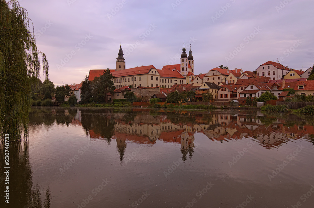 Scenic view of Telc castle, lake, Name of Jesus Church and tower of the Church of St. Jakub. Buildings are reflected in the water. Early morning landscape. A UNESCO World Heritage Site