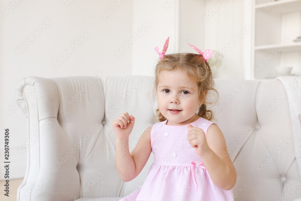 Cute little girl in pink dress looking at camera and gesturing with hands while sitting on comfortable couch in stylish room Cute little girl in pink dress looking at camera and gesturing with hands while sitting on comfortable couch in stylish room