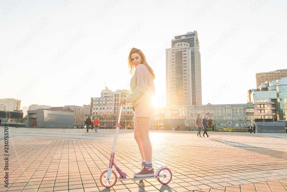 Cute girl student in pink clothes rides a kick scooter along the streets of the city at sunset. Girl rides the city on ecological transport, looks into the camera and smiles. Scooter Concept. Cute girl student in pink clothes rides a kick scooter along the streets of the city at sunset. Girl rides the city on ecological transport, looks into the camera and smiles. Scooter Concept.