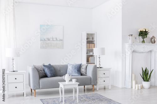 Table on carpet in front of grey settee in white living room interior with lamps and poster. Real photo