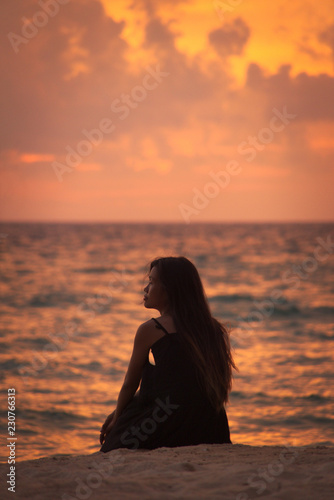Young asian Vietnamese woman sitting on sand silhouetted on beach posing with sunset sky and sea in background
