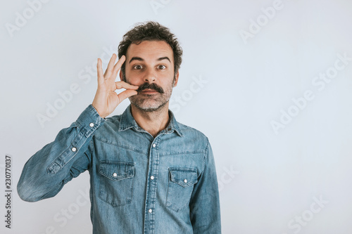 Funny portrait of brown, smiling, handsome man touching his mustache. Jeans shirt. Gray background. photo