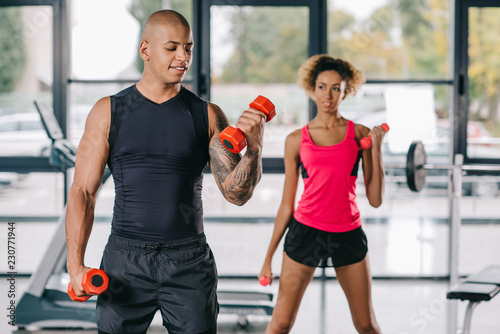 smiling african american male trainer training young woman to exercising with dumbbells at gym