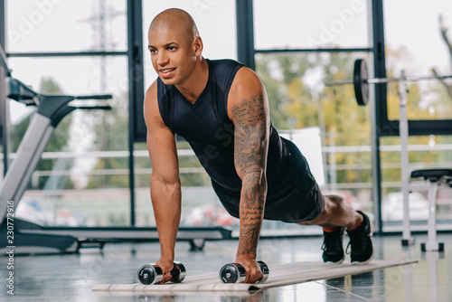 handsome african american sportsman with tattooed hand doing push ups with dumbbells on fitness mat at gym