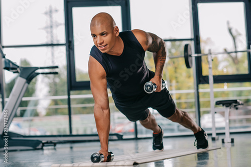 smiling muscular african american sportsman exercising with dumbbells on fitness mat at gym