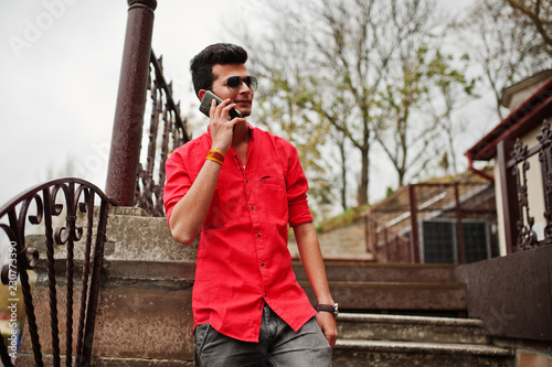Indian man at red shirt and sunglasses posed outdoor and speaking on phone. photo
