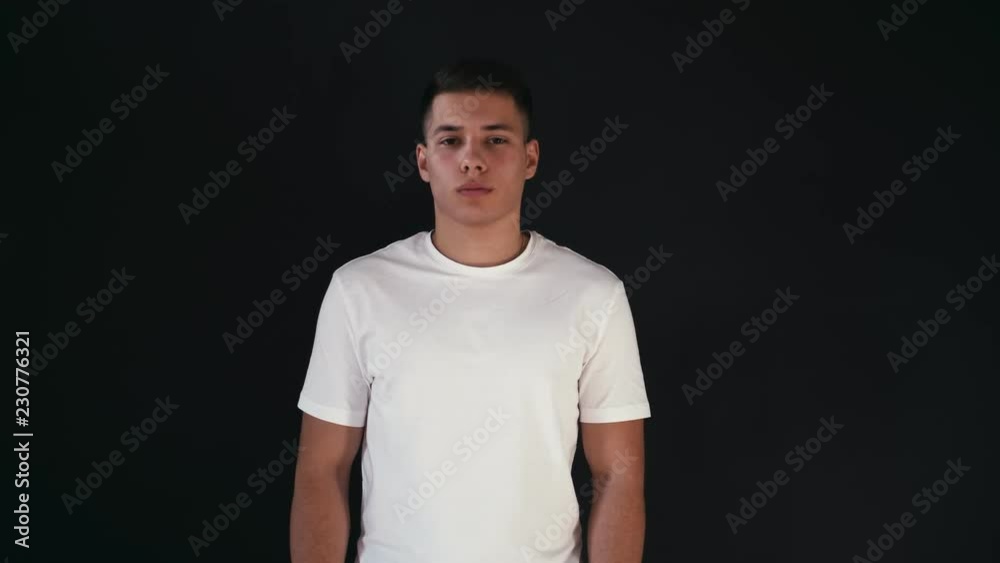 Attractive and confident young man stands in white shirt and poses on camera. He lifts up hands and crosses them. Guy looks very confident and good. Isolated on black background. Attractive and confident young man stands in white shirt and poses on camera. He lifts up hands and crosses them. Guy looks very confident and good. Isolated on black background.