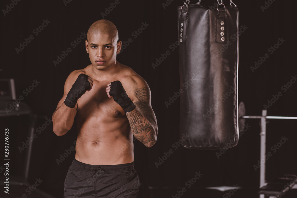 Serious African American boxer posing in fight position near punching bag at gym Serious African American boxer posing in fight position near punching bag at gym