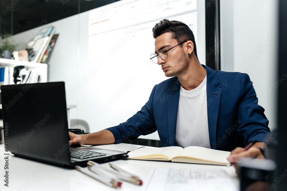 Dark-haired young architect dressed in a blue jacket works on the laptop in the office Dark-haired young architect dressed in a blue jacket works on the laptop in the office