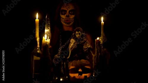 Halloween witch with skull makeup makes voodoo holds knife and wispering spell magic pumpkin chains and candles photo