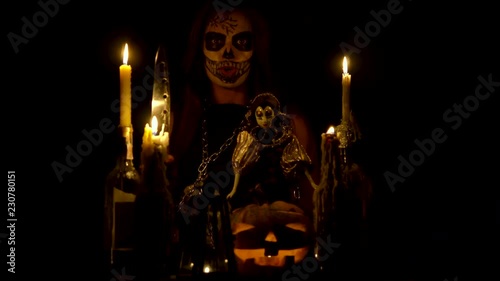 Halloween witch with skull makeup makes voodoo holds knife and wispering spell magic pumpkin chains and candles