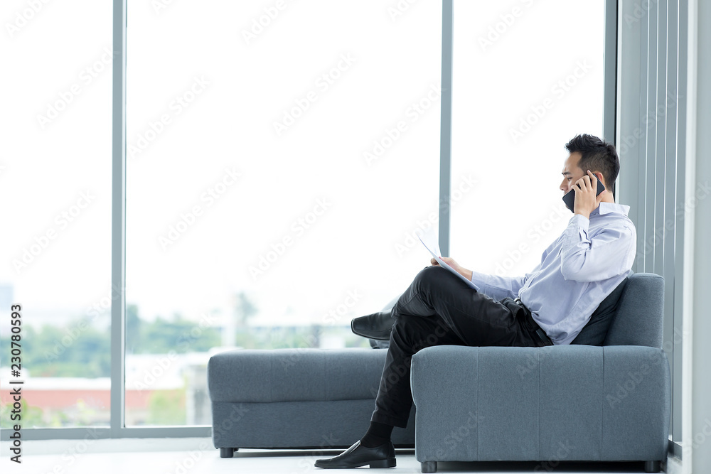 Young Asian businessman using mobile smartphone sitting on sofa serious and have ducuments at hand in the modern office with big windows Young Asian businessman using mobile smartphone sitting on sofa serious and have ducuments at hand in the modern office with big windows