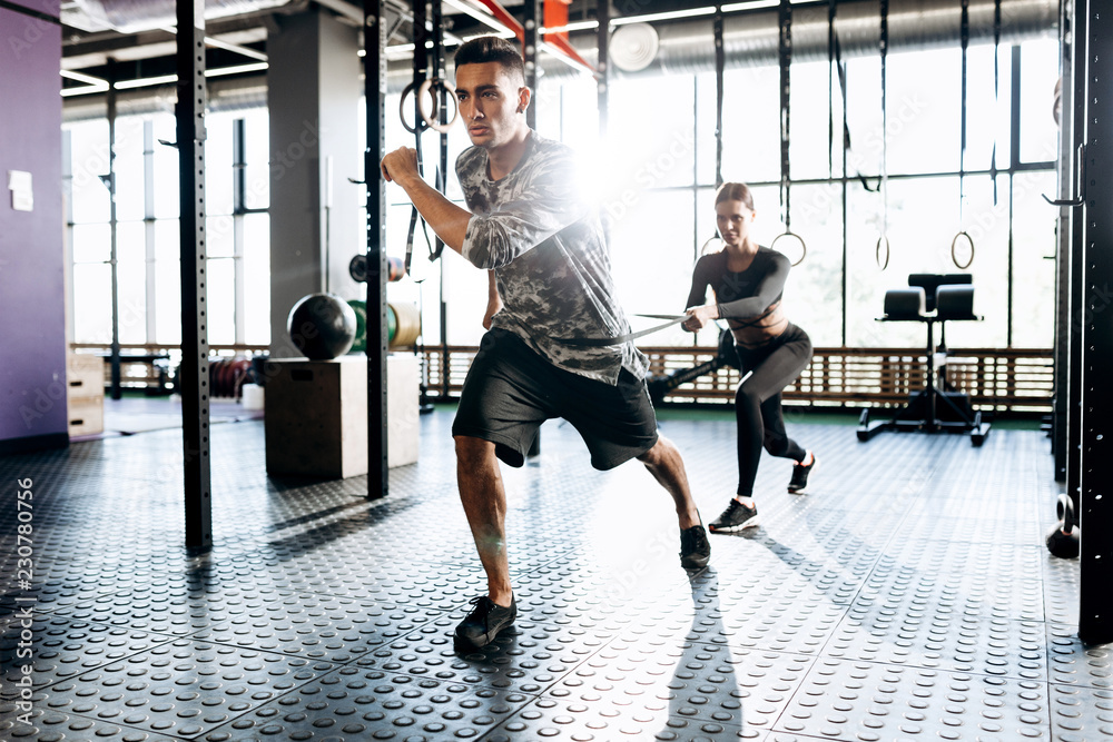 Obraz premium Young athletic man and slim girl are doing sport exercises with special equipment such as power band in the gym