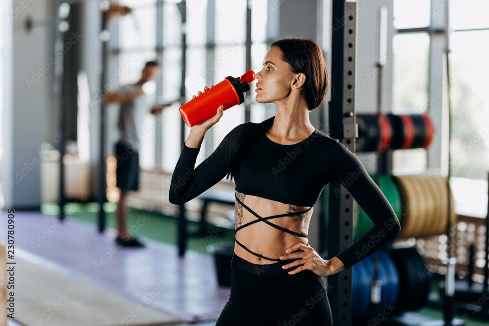 Fototapeta Slim dark-haired girl dressed in black sportswear drinks water in the gym near the sport equipment
