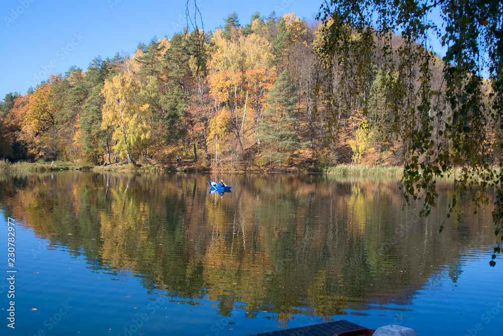 Beautiful autumnal forest is mirrored in the calm waters of a lake, with a fishermen in a blue boat Beautiful autumnal forest is mirrored in the calm waters of a lake, with a fishermen in a blue boat