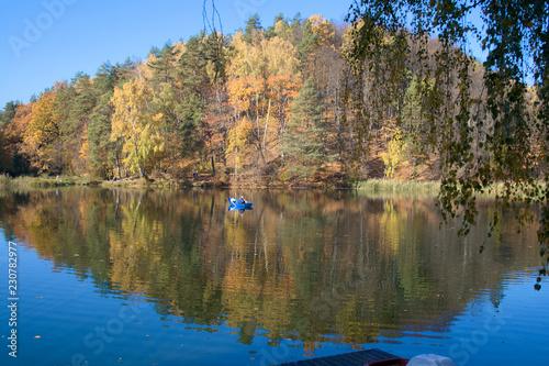 Fototapeta Naklejka Na Ścianę i Meble -  Beautiful autumnal forest is mirrored in the calm waters of a lake, with a fishermen in a blue boat 