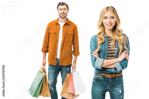 beautiful smiling woman posing with crossed arms while her boyfriend standing behind with shopping bags isolated on white