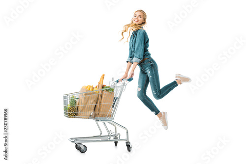 positive beautiful girl jumping near shopping trolley cart with products in paper bags isolated on white photo