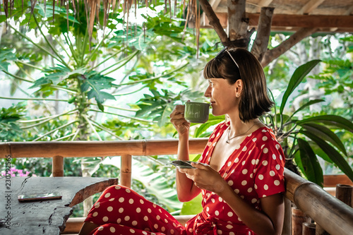 Young woman in red dress drinking a coffee in a tropical restaurant on Bali island. Travel, jungle, rainforest.