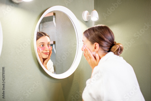 Young woman in bathrobe taking care of herself looking into the mirror with patches under eyes in the bathroom