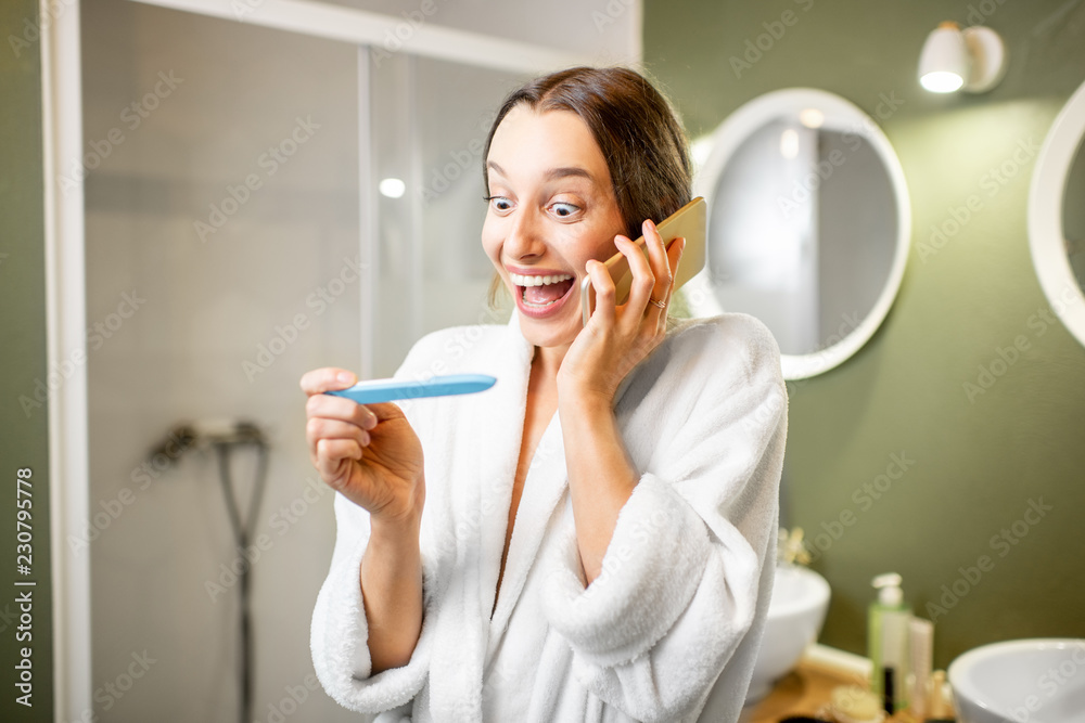 Happy woman in bathrobe excited with a pregnancy test result calling with phone in the bathroom Happy woman in bathrobe excited with a pregnancy test result calling with phone in the bathroom