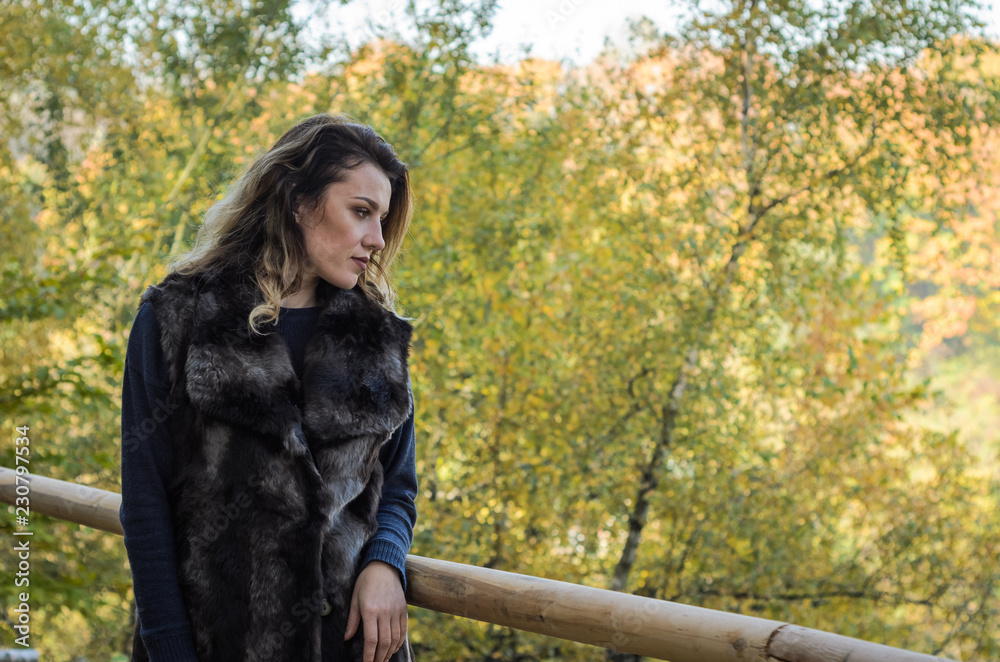 Young beautiful girl with long hair, in a fur jacket, stands on a wooden railing on a bright sunny day while walking in the autumn park Young beautiful girl with long hair, in a fur jacket, stands on a wooden railing on a bright sunny day while walking in the autumn park