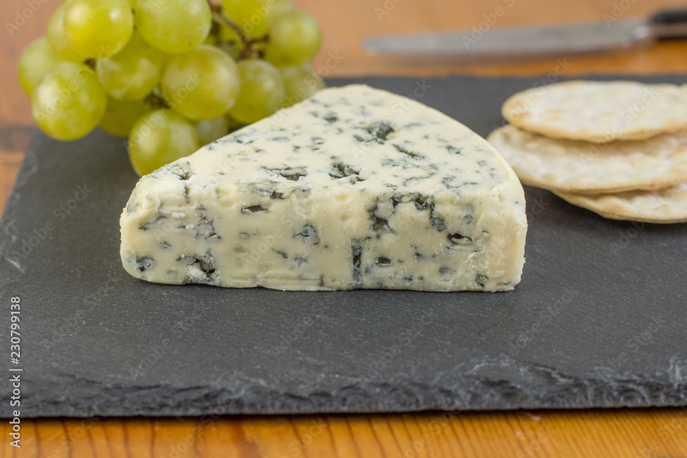 Blue cheese wedge on slate board close up with defocused green grapes and crackers in background Blue cheese wedge on slate board close up with defocused green grapes and crackers in background