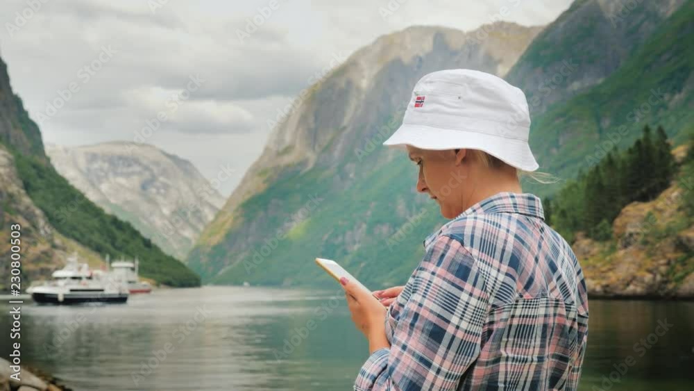 A tourist uses a smartphone on the background of the picturesque Norwegian fjord, in the distance a cruise ship is visible. Journey to Scandinavia A tourist uses a smartphone on the background of the picturesque Norwegian fjord, in the distance a cruise ship is visible. Journey to Scandinavia