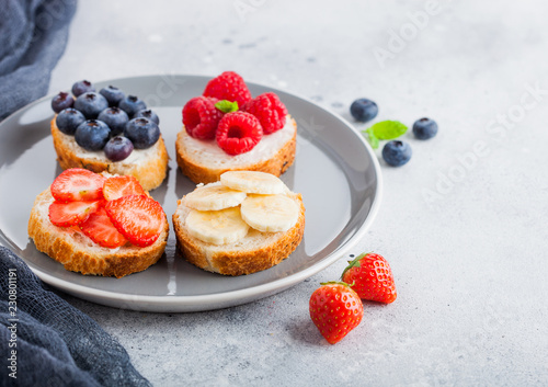 Fresh healthy mini sandwiches with cream cheese, fruits and berries in grey plate with cloth. Strawberries, blueberries, bananas and raspberries on stone kitchen table background.Top view.
