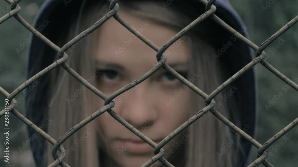 Portrait of moody and sad blonde caucasian girl behind the iron fence. Young woman behind metal fence grid jail locked