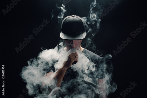 Vape man. Portrait of a handsome young white guy in a modern black cap vaping and letting off puffs of steam from an electronic cigarette  photo