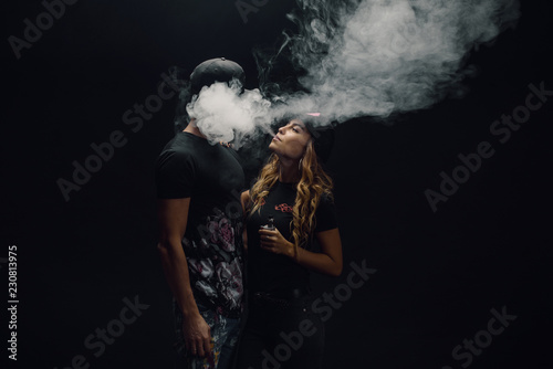 Couple vaping. Young man and woman blowing smoke to join it in one cloud at black studio background. Relationship and vape addiction concept with copy space photo