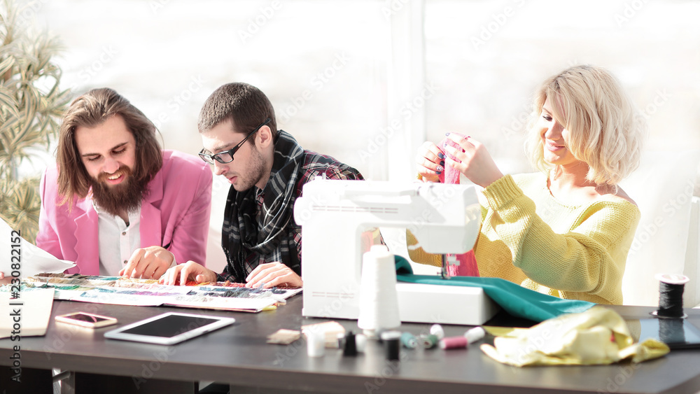 Smiling tailor businesswoman sitting at sewing machine and making handmade clothing while marketing assistant working on new projec Smiling tailor businesswoman sitting at sewing machine and making handmade clothing while marketing assistant working on new projec