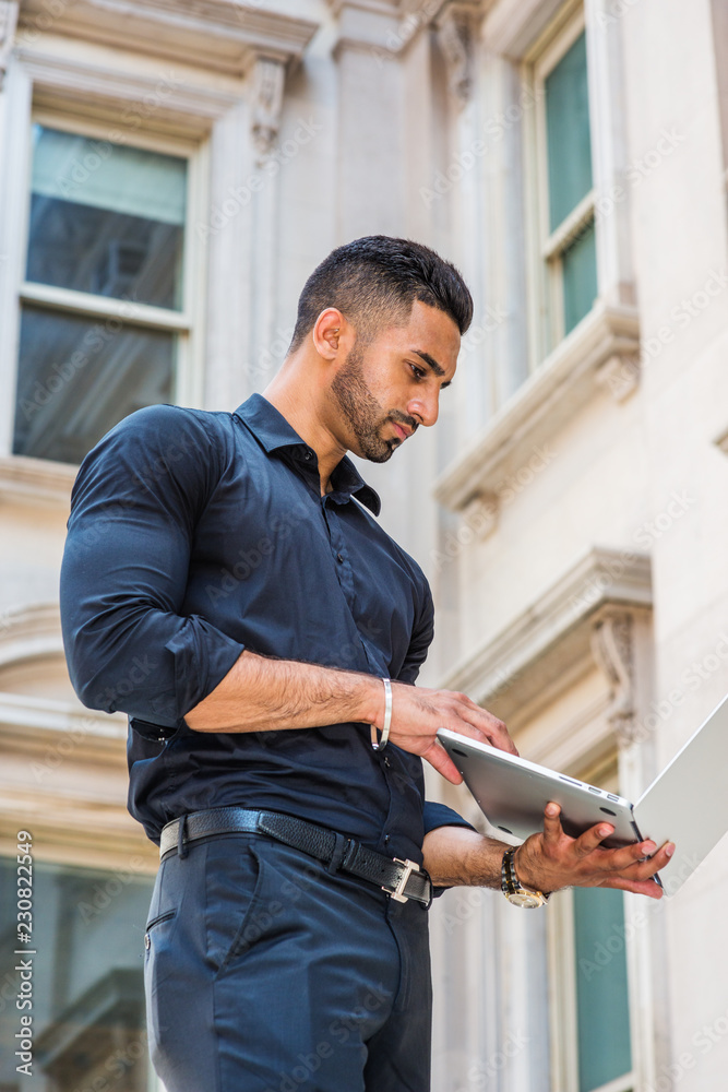 Way to Success. Young East Indian American Businessman with beard working in New York, wearing black shirt, holding laptop computer, standing outside old style office building, looking down, reading.. Way to Success. Young East Indian American Businessman with beard working in New York, wearing black shirt, holding laptop computer, standing outside old style office building, looking down, reading..
